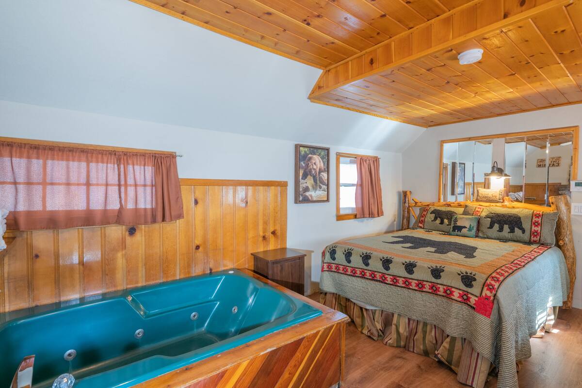 A room with a wooden ceiling, a large bed with a bear-themed bedspread, a wooden headboard, a window with a brown curtain, and a wooden floor.