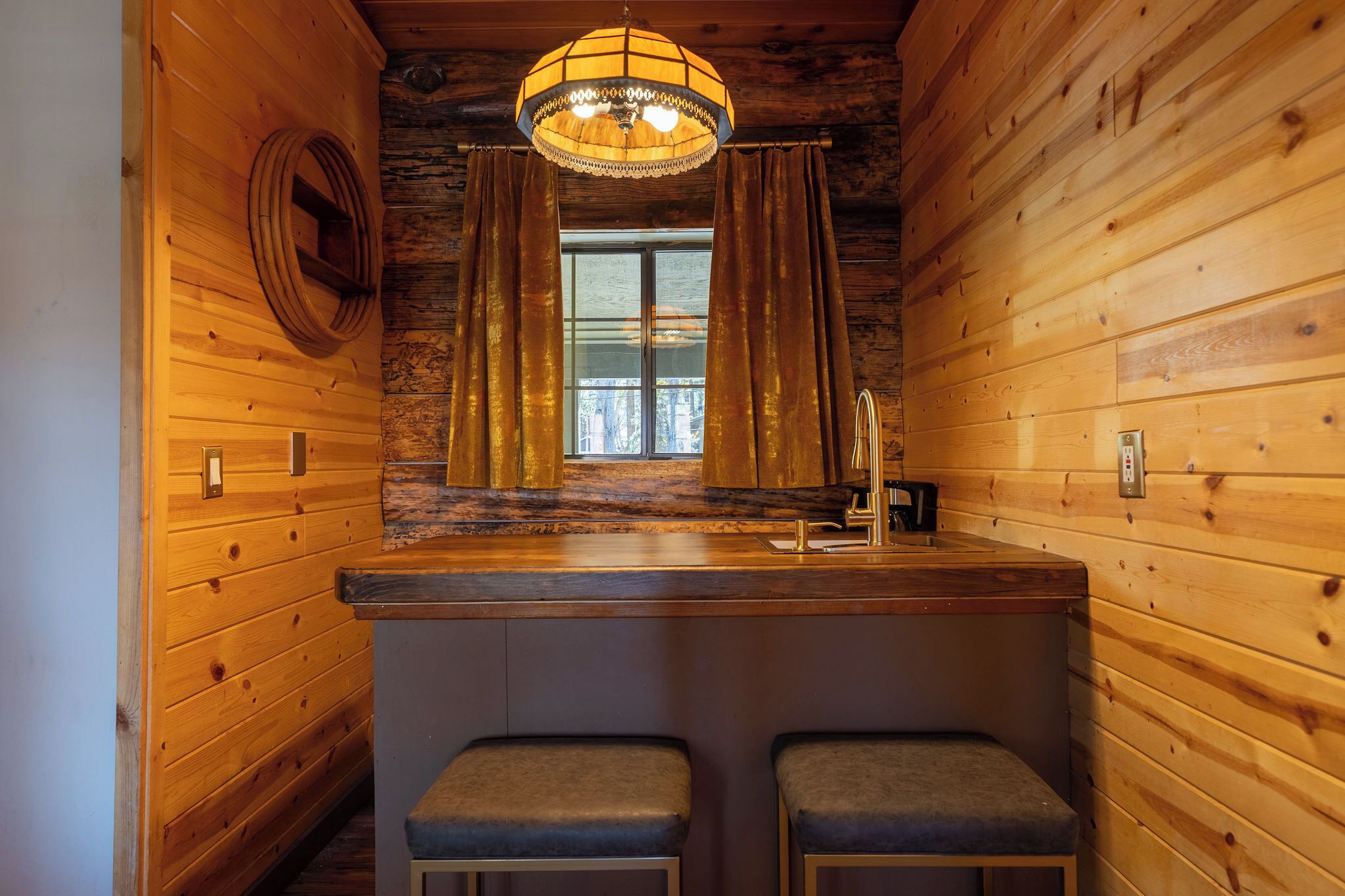 A wooden cabin interior with a bar area, two stools, a window with curtains, and a hanging light fixture.