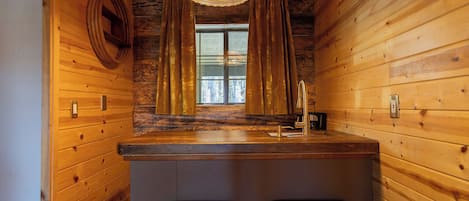 A wooden cabin interior with a bar area, two stools, a window with curtains, and a hanging light fixture.
