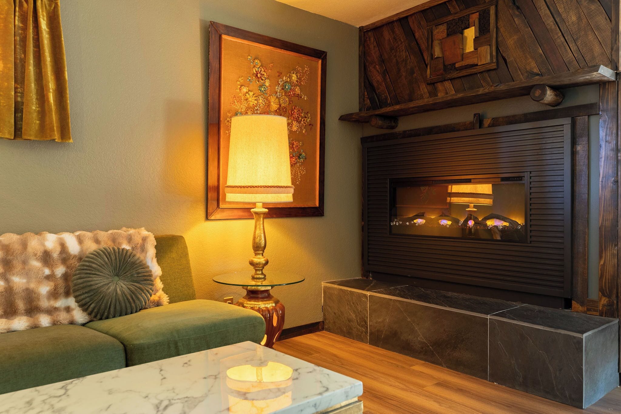 A cozy living room with a green sofa, a marble coffee table, a lamp, a fireplace, and a framed floral artwork on the wall.
