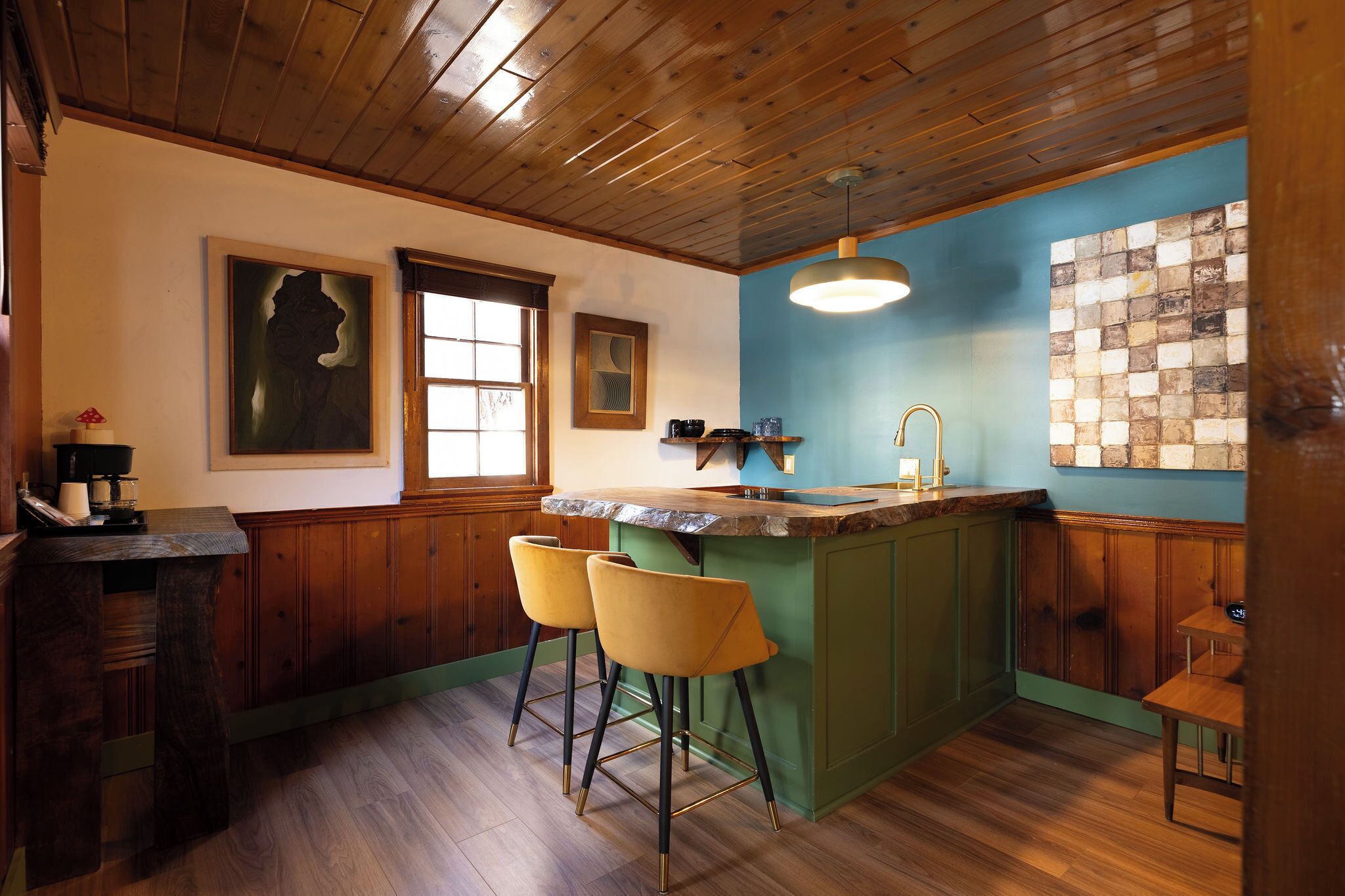 Een keuken met houten kastjes, een groen kookeiland met barstoelen, een raam en een blauwe accentmuur.