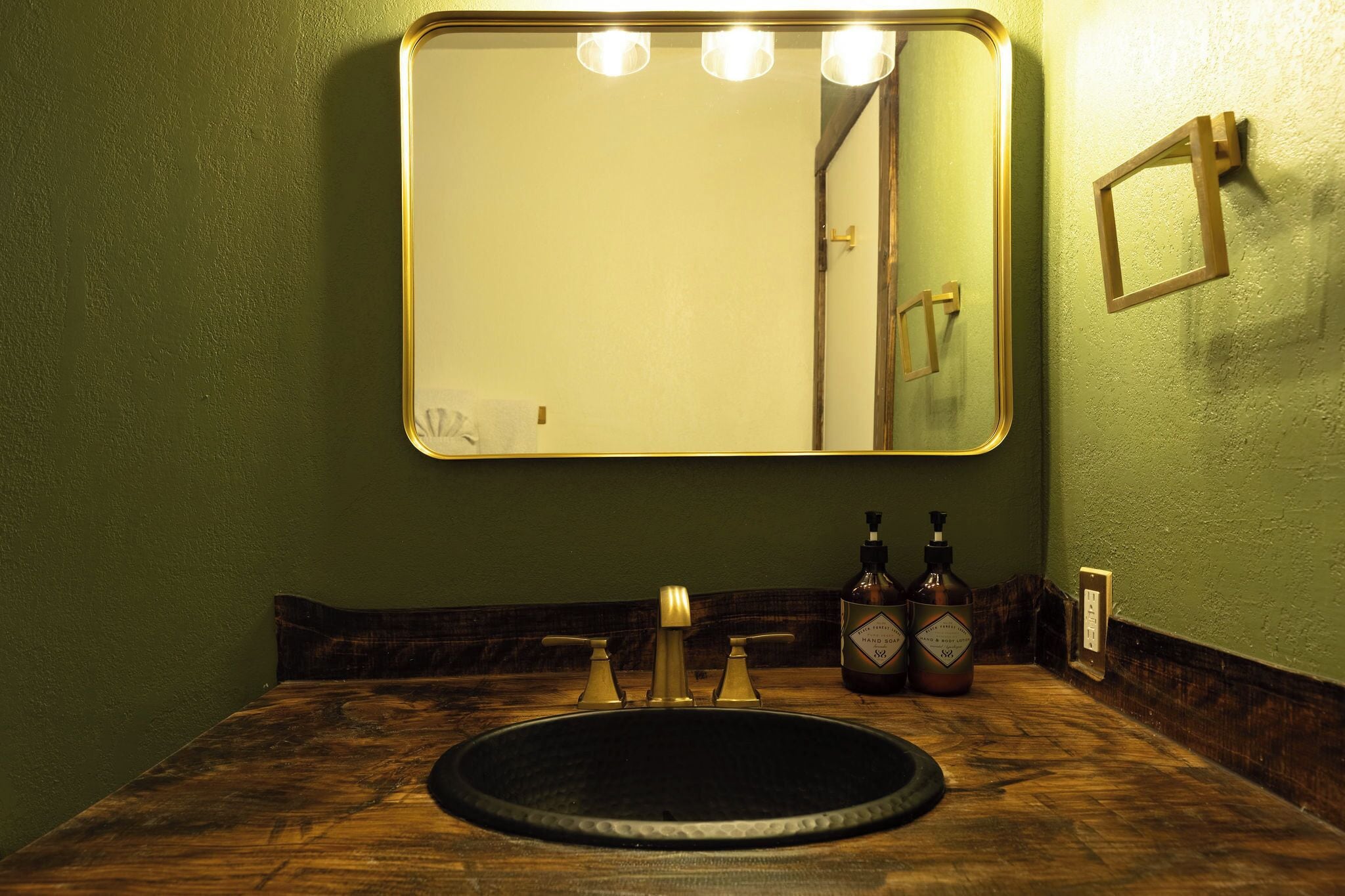 Une salle de bain avec un meuble-lavabo en bois, un lavabo rond et un miroir avec éclairage intégré. On peut voir deux bouteilles posées sur le comptoir.