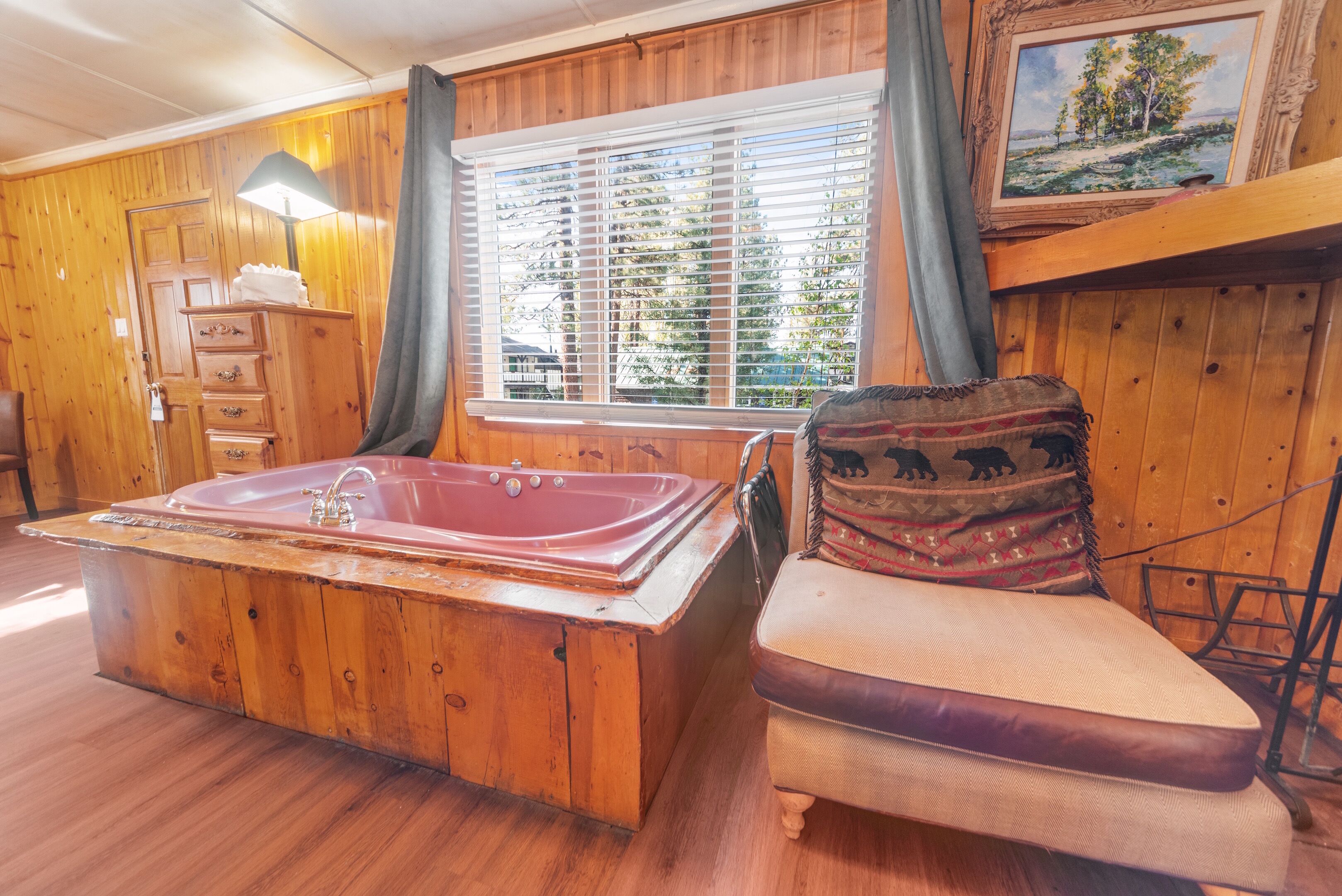 A cabin interior with a wooden floor, a large wooden bathtub, a cushioned chair, a wooden dresser, a lamp, and a window with blinds.