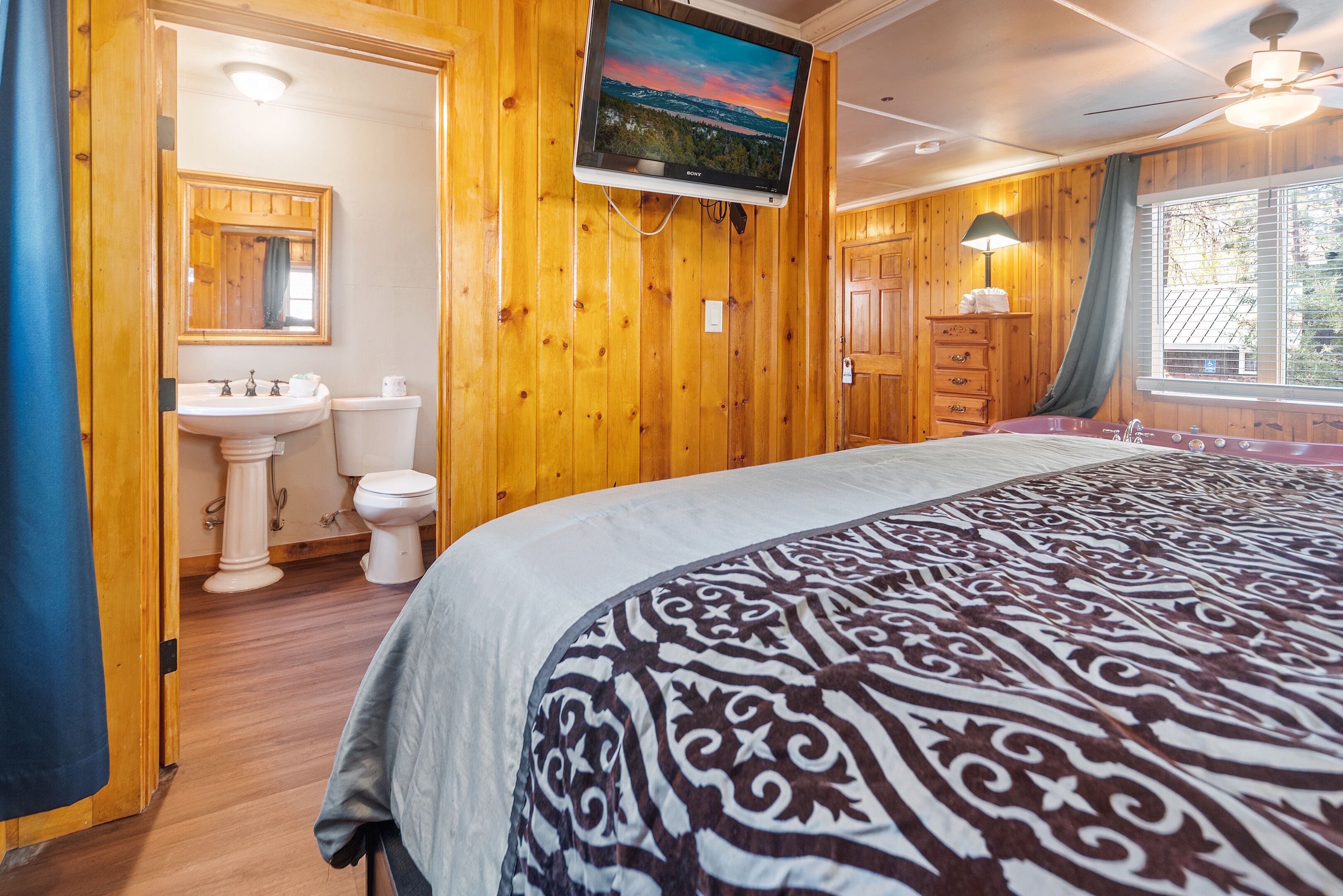 Habitación con paneles de madera, una cama, televisor montado, un lavamanos y una ventana con cortinas.