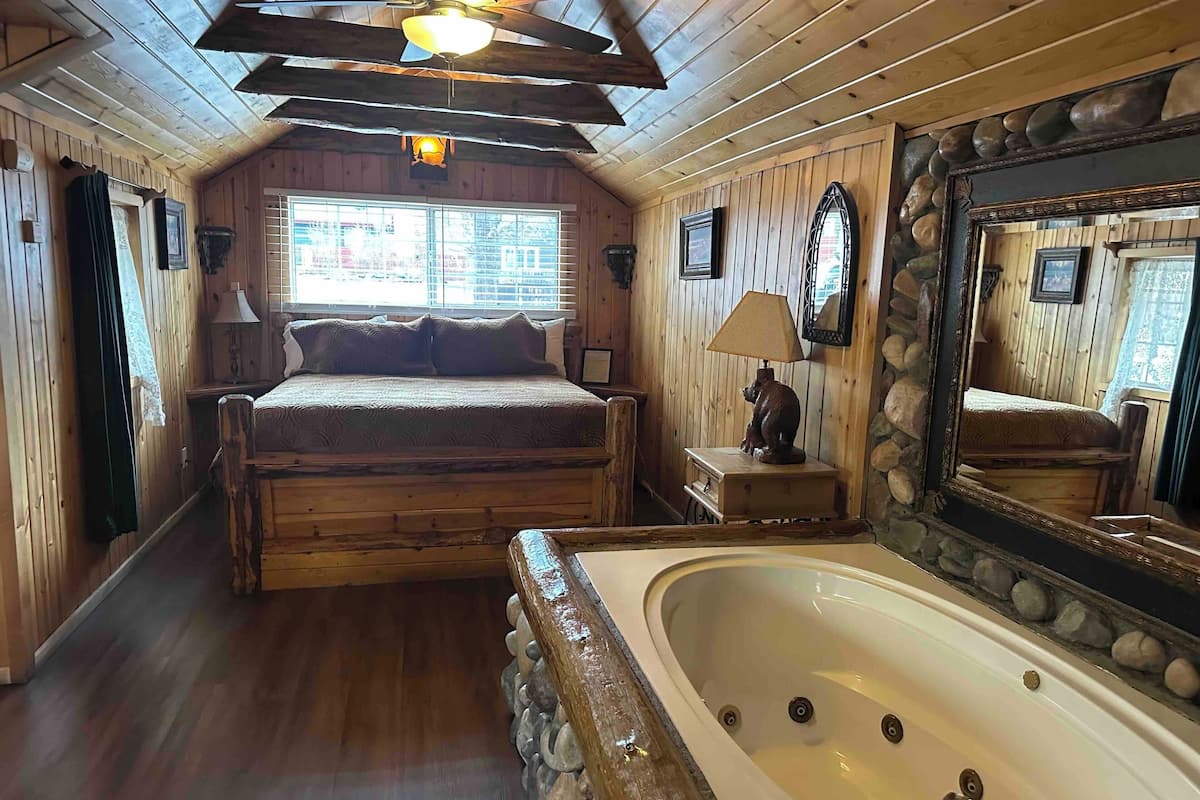 A wooden cabin room with a bed, a large mirror, a bathtub, and a ceiling fan.
