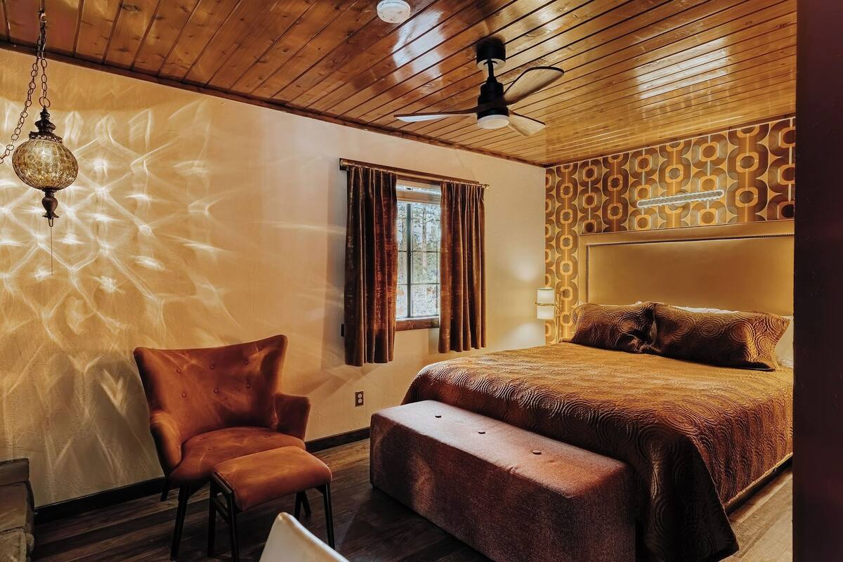 A bedroom with a wooden ceiling, a bed with a patterned headboard, a hanging lamp, and a window with curtains.