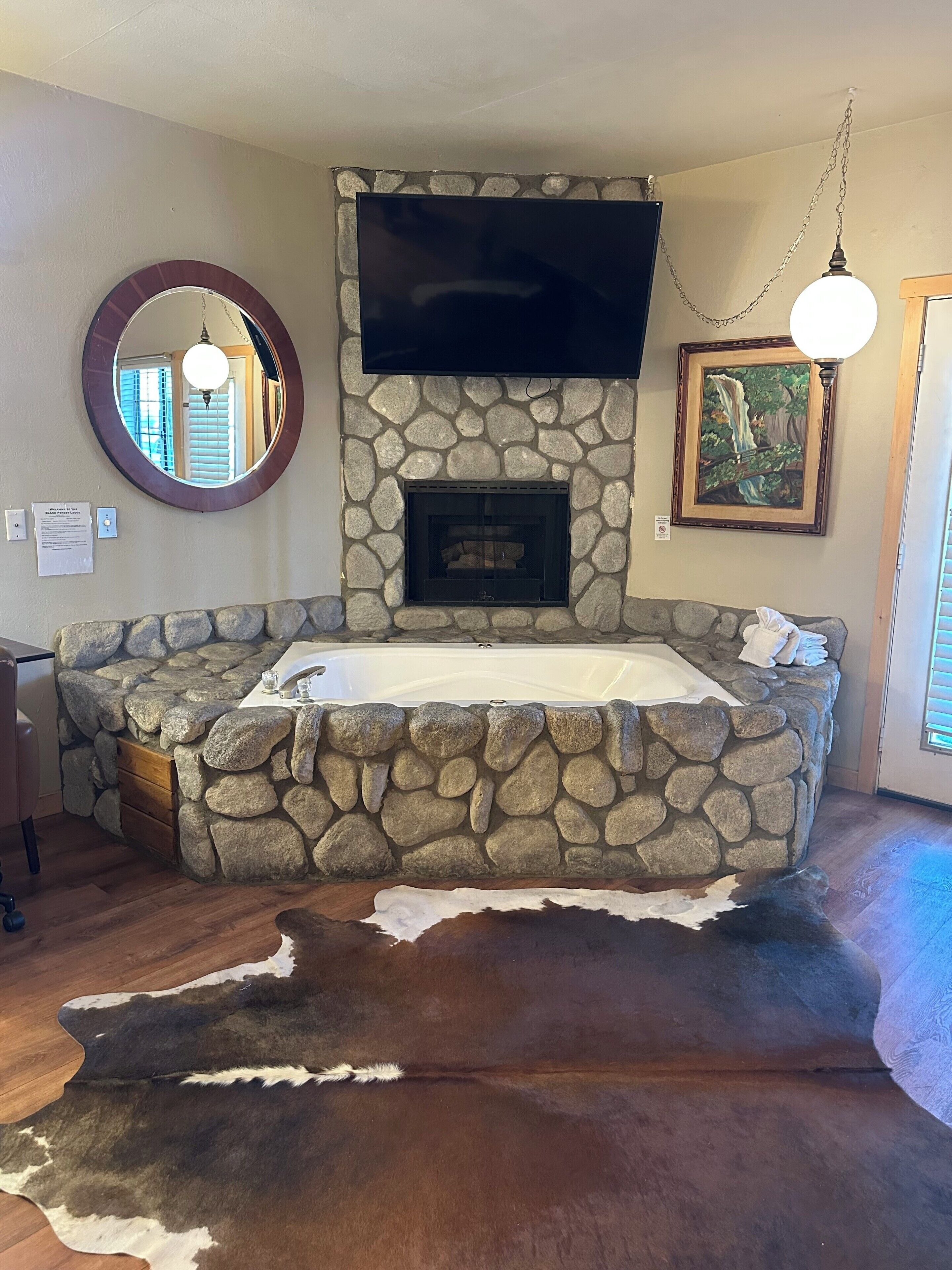 A stone fireplace with a flat-screen TV above it, a round mirror on the wall, and a framed painting.