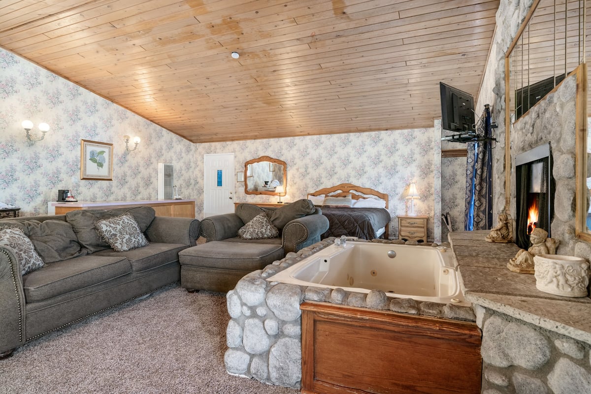 A cozy living room with a stone fireplace, a large stone bathtub, and a wooden ceiling.