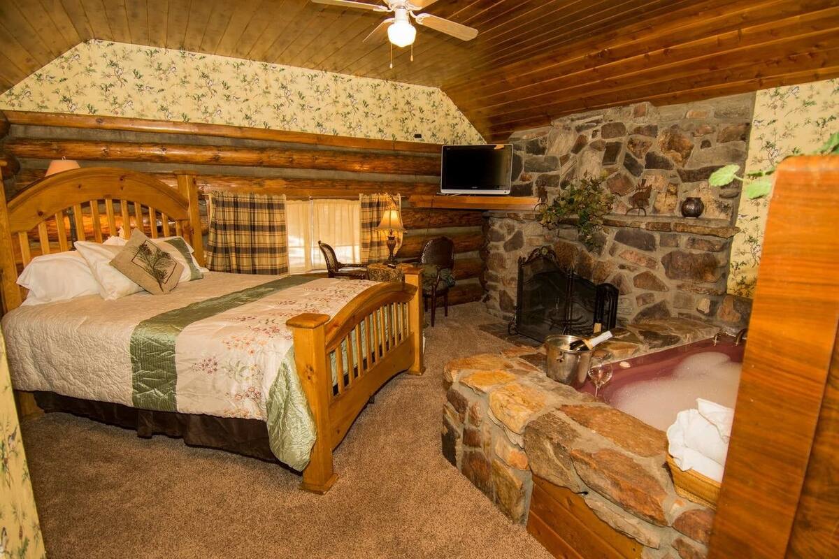 A cozy log cabin bedroom with a bed, a stone fireplace, a stone bench, and a television mounted on the wall.