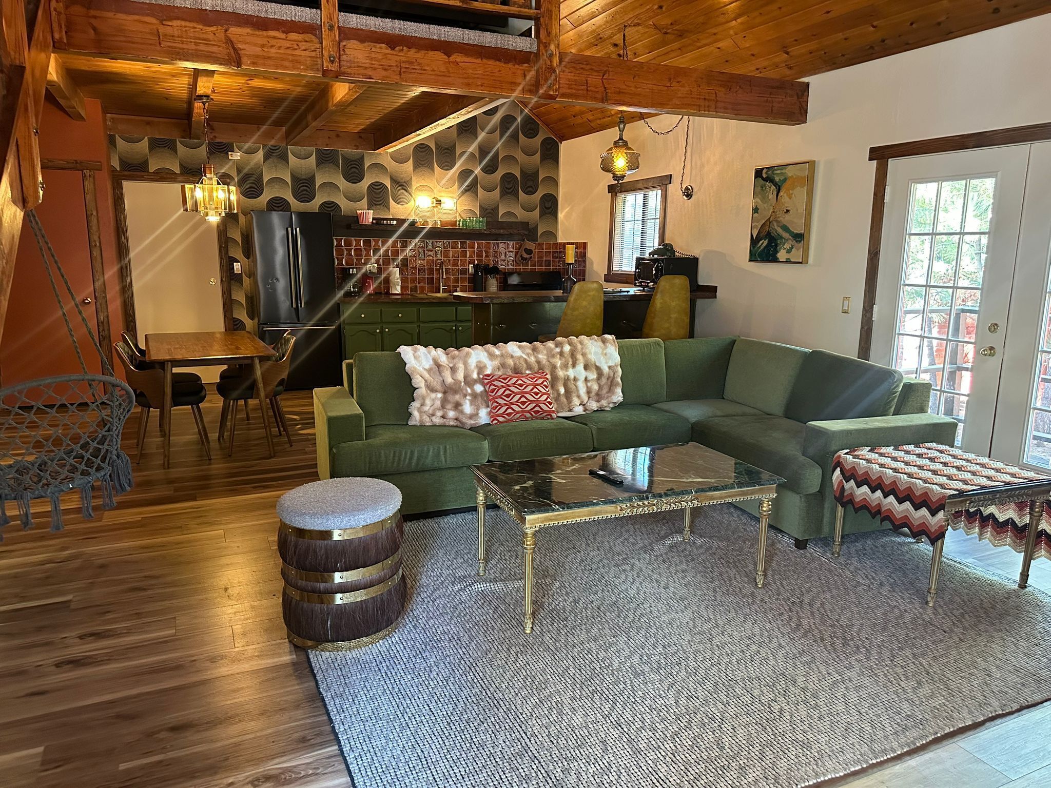 A cozy living room with a green sofa, a coffee table, and a hanging chair. A kitchen with a wooden ceiling and a dining area with a table and chairs are visible.