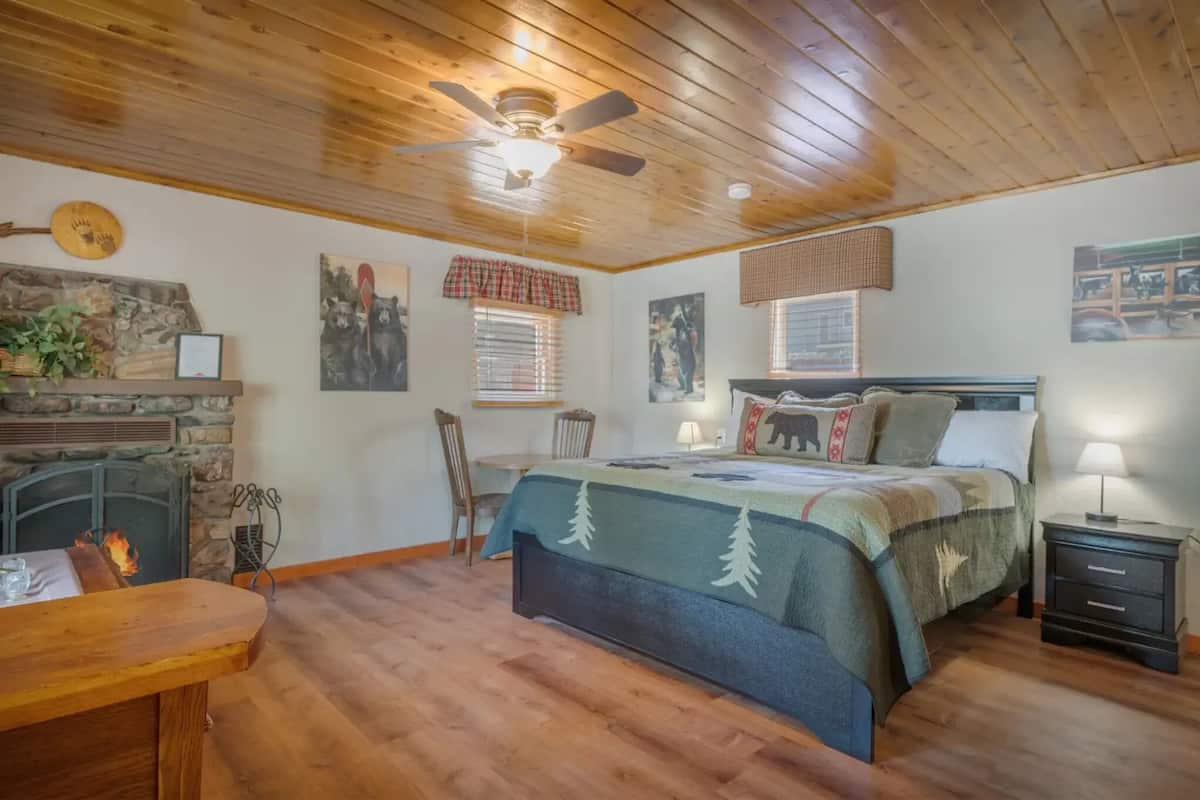 A cozy bedroom with a stone fireplace, a bed with forest-themed bedding, a ceiling fan, and a small table with a lamp.
