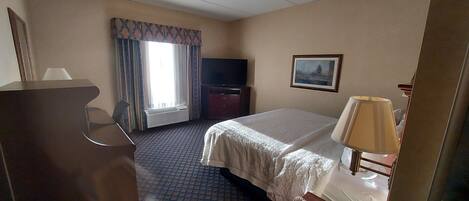 Una habitación de hotel con cama, escritorio, televisión, ventana con cortinas y un cuadro en la pared.
