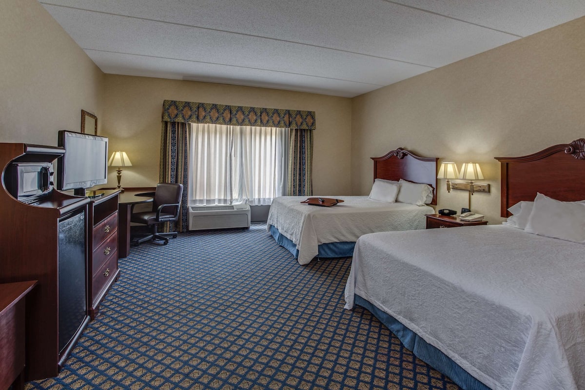 A hotel room with two beds, a desk with a television, a chair, and a window with curtains.