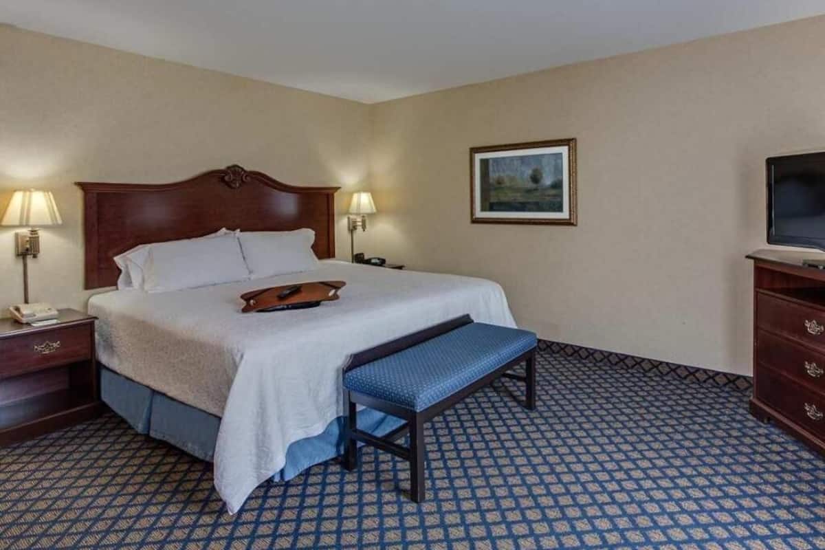 A hotel room with a large bed, bedside lamps, a bench, a dresser, and a television.
