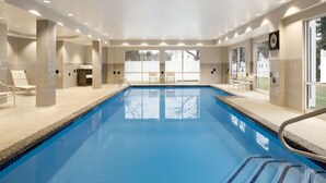 A large indoor swimming pool with clear blue water, surrounded by beige tiles and columns. There are chairs and a small table near the pool, and large windows on one side. - Hampton Inn Dover (Dover)