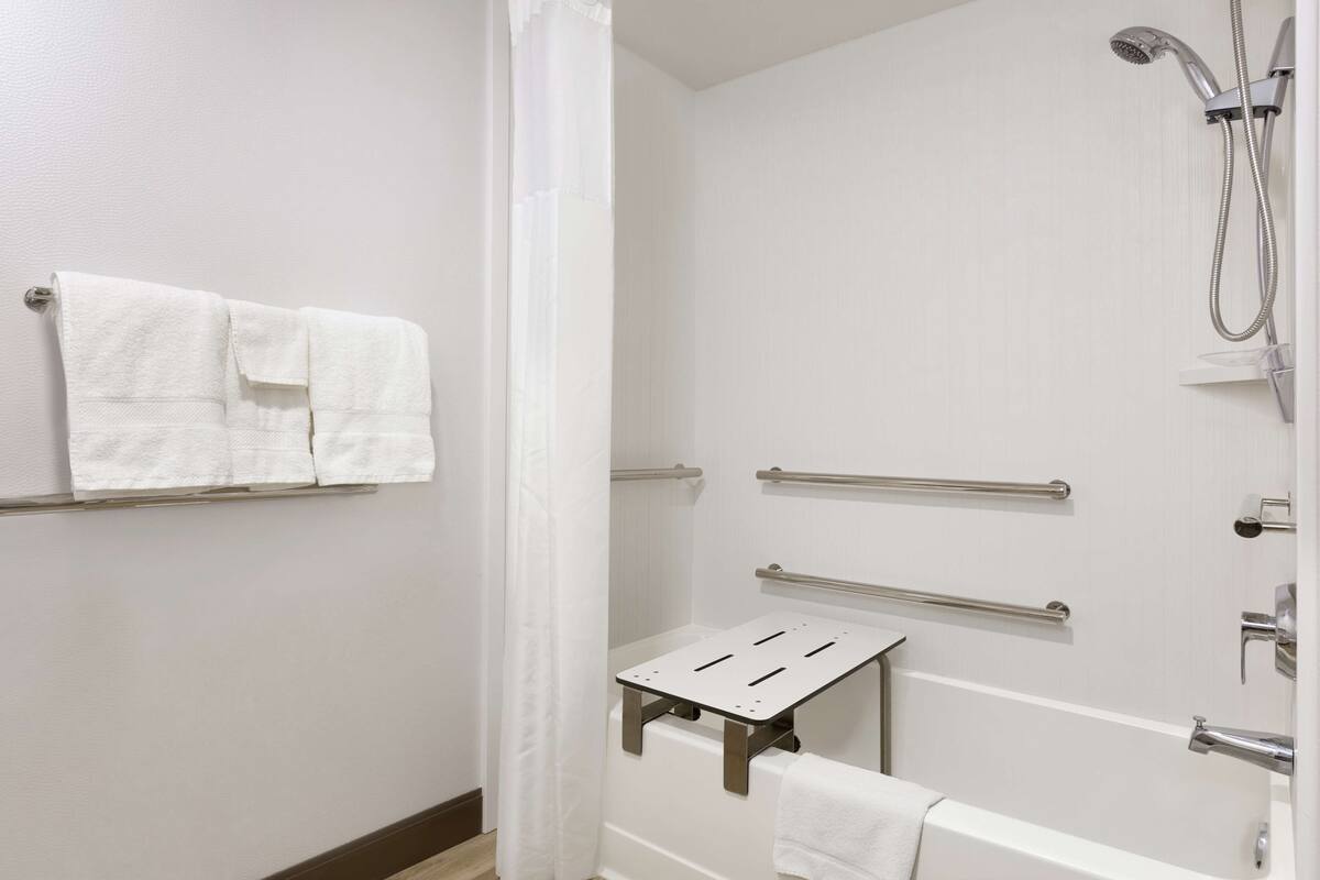 A bathroom with a shower, a bathtub, and a small table. There are handrails and a towel rack.