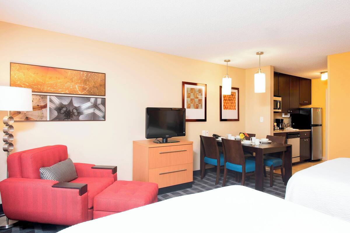 A hotel room with a red sofa, a TV on a wooden cabinet, a dining table with chairs, and a kitchenette in the background.