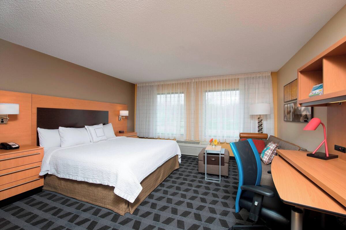 A hotel room with a large bed, a desk with a chair, a small table with a lamp, and a nightstand with a phone.