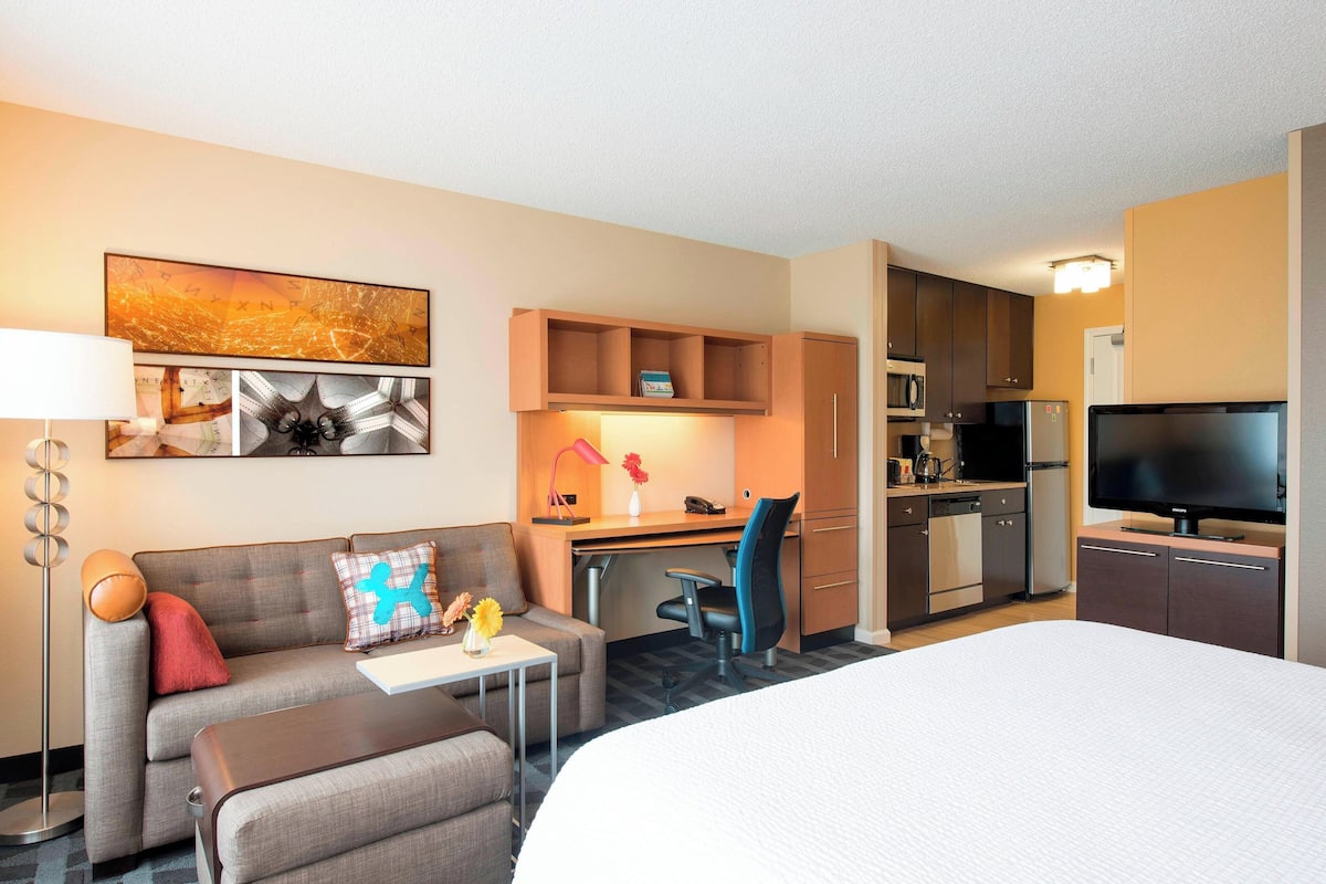 A hotel room with a bed, a sofa, a desk, a chair, a TV, a kitchen area with a microwave, refrigerator, and a flat-screen TV.