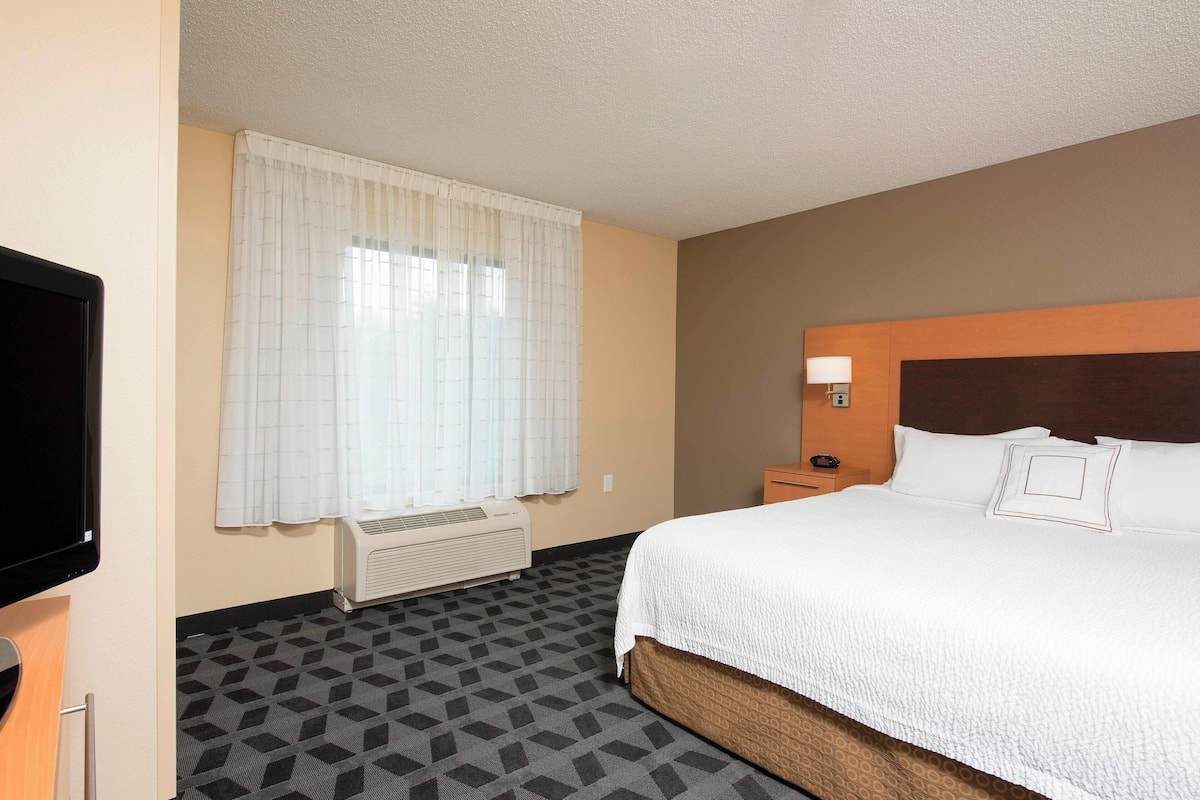 A hotel room with a large bed, a flat-screen TV, a window with curtains, a heater, and a nightstand with a lamp.
