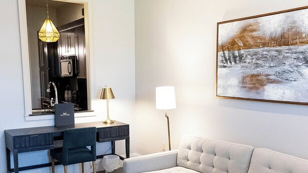 A modern hotel room with a sofa, a wooden coffee table, a desk with a chair, a lamp, and a painting on the wall. - Carriage House Hotel (Lafayette)