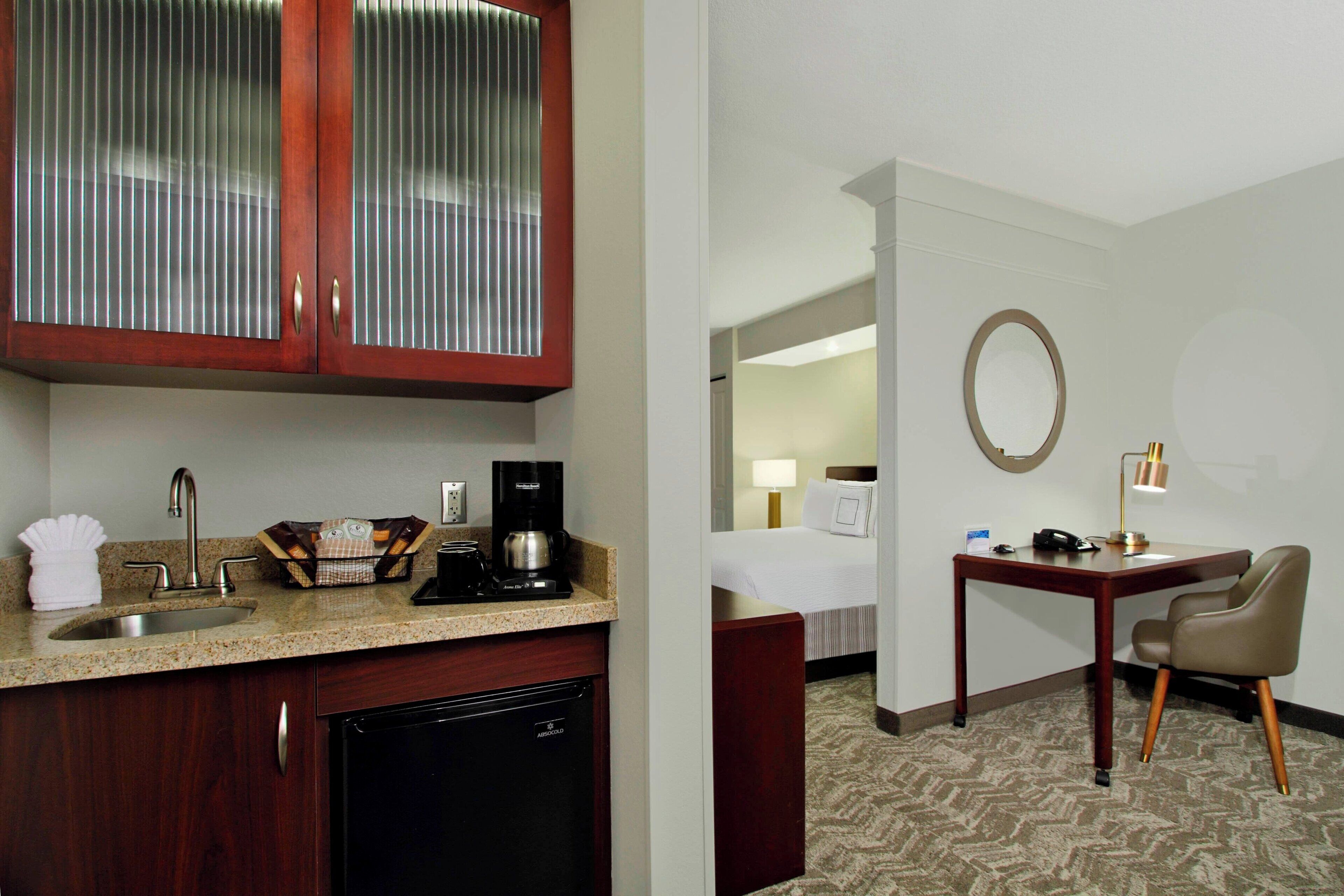 Habitación de hotel con cocina que incluye un lavabo, cafetera y armarios. Hay un escritorio con silla y un espejo redondo en la pared.