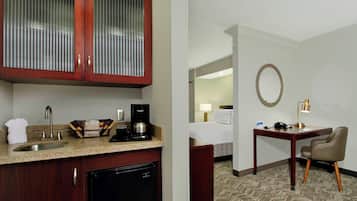 Habitación de hotel con cocina que incluye un lavabo, cafetera y armarios. Hay un escritorio con silla y un espejo redondo en la pared.