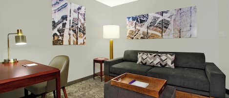 A modern hotel room with a gray sofa, a wooden side table, a desk with a lamp, and two abstract paintings on the walls.