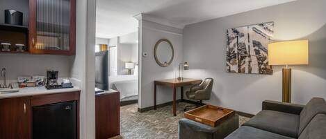 A hotel room with a kitchenette, a sofa, a desk, a chair, a lamp, a mirror, and a painting on the wall.