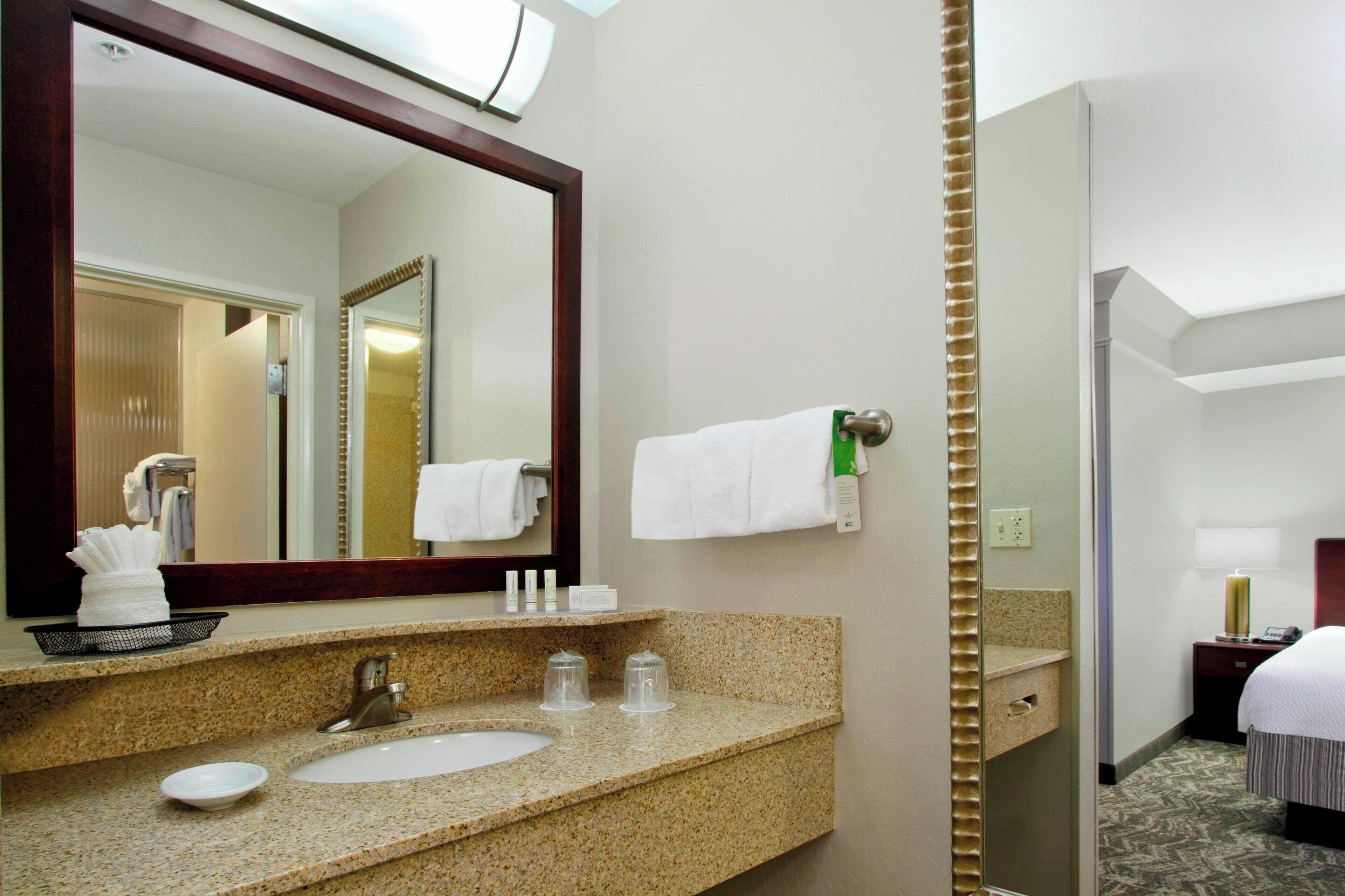 Un bagno d'hotel con un piano di lavoro in granito, lavabo, specchio, portasciuga e un letto nella stanza adiacente.