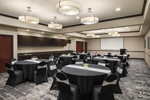Un salón de conferencias con varias mesas, sillas negras adornadas con cintas blancas y candelabros colgantes.