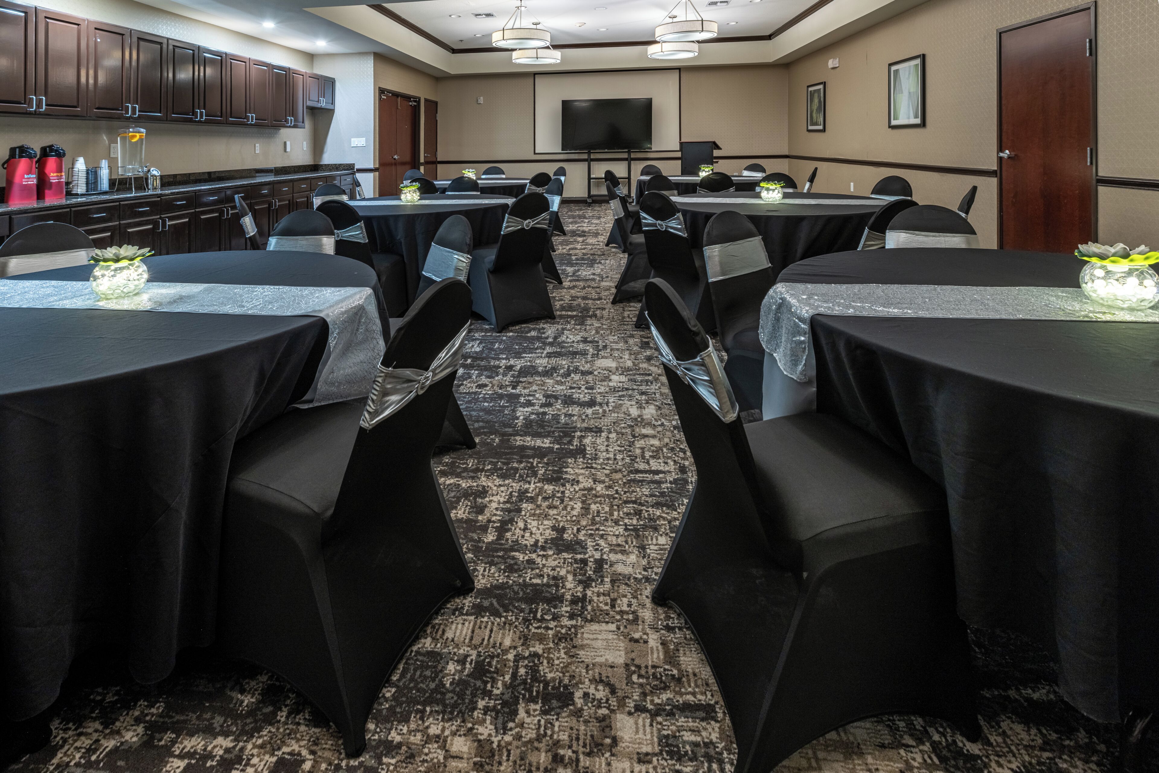 A conference room with multiple tables and chairs, equipped with a projector screen and a television.