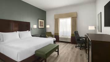 A hotel room with a large bed, a desk, a chair, a bench, a window with curtains, and a wall-mounted lamp.