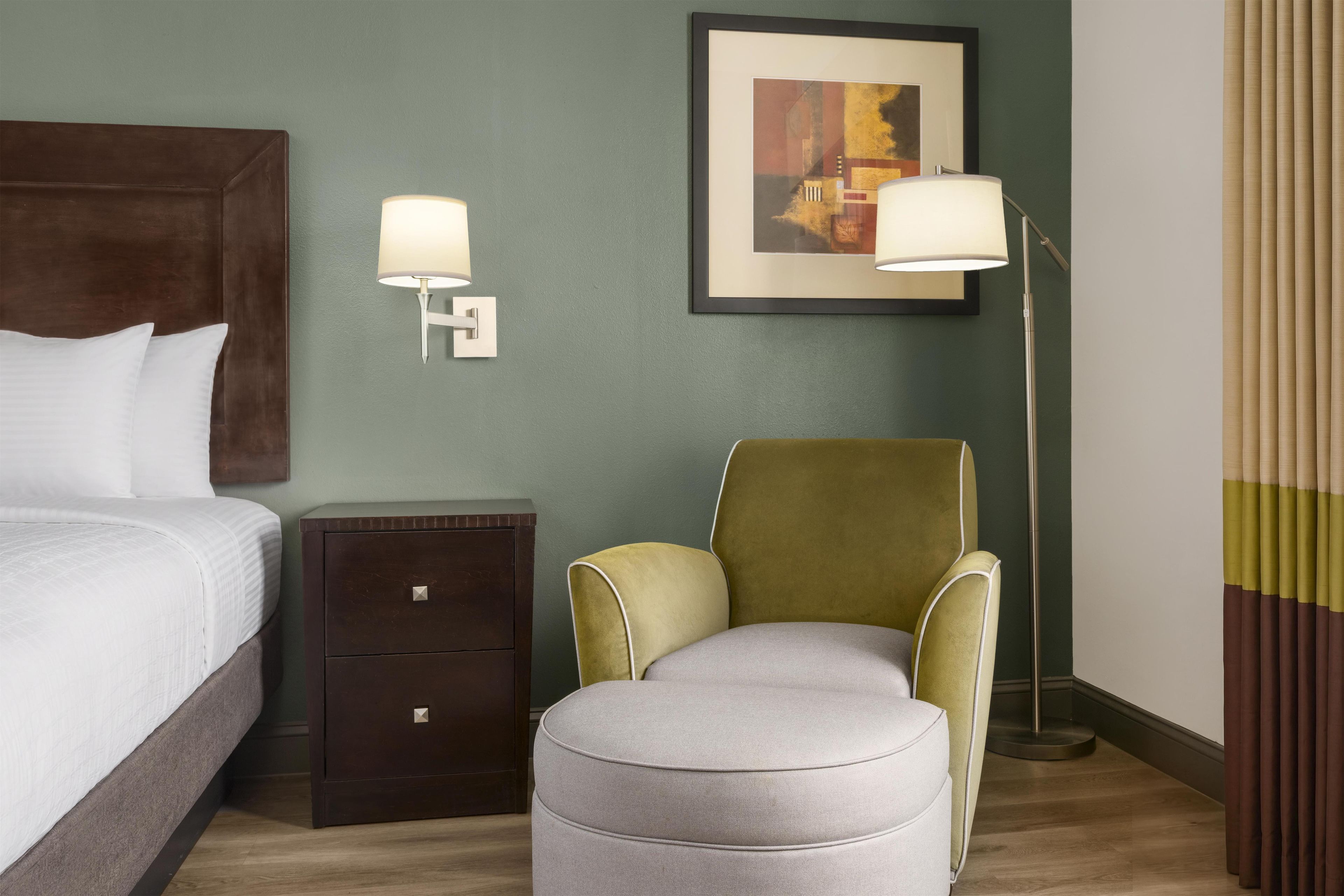 Ein Hotelzimmer mit Bett, Nachttisch, Lampe, Stuhl und einem gerahmten Kunstwerk an der Wand.