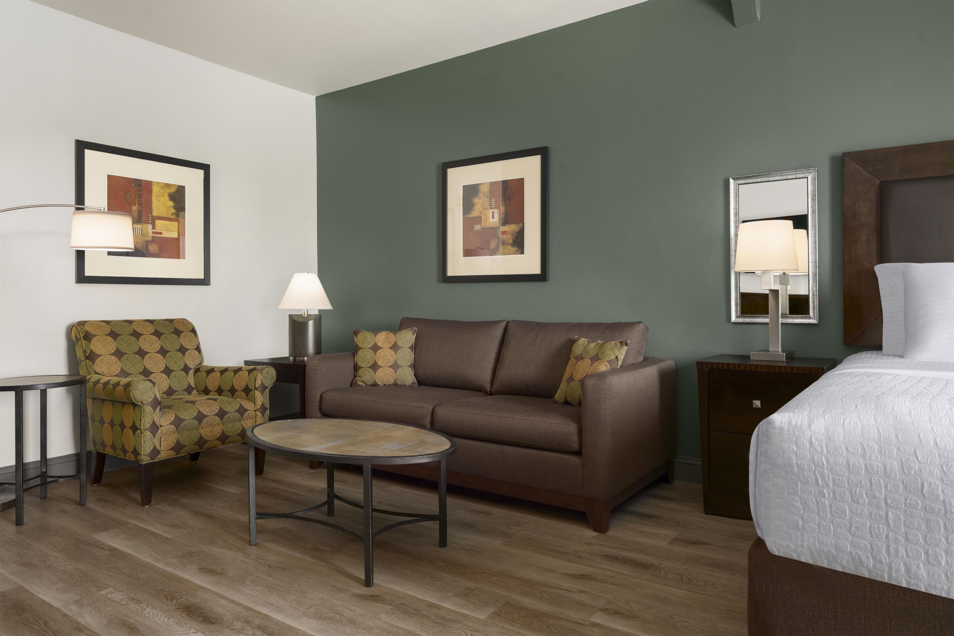Une chambre d’hôtel avec un canapé marron, une table basse ronde, un fauteuil à motifs, un lit recouvert d’une couette blanche et deux tableaux abstraits encadrés accrochés aux murs.