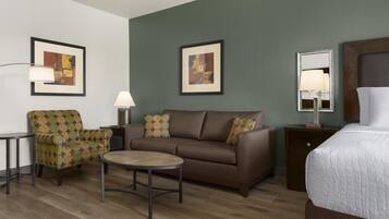 Une chambre d’hôtel avec un canapé marron, une table basse ronde, un fauteuil à motifs, un lit recouvert d’une couette blanche et deux tableaux abstraits encadrés accrochés aux murs.