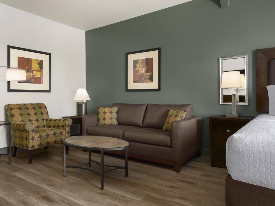 Une chambre d’hôtel avec un canapé marron, une table basse ronde, un fauteuil à motifs, un lit recouvert d’une couette blanche et deux tableaux abstraits encadrés accrochés aux murs.