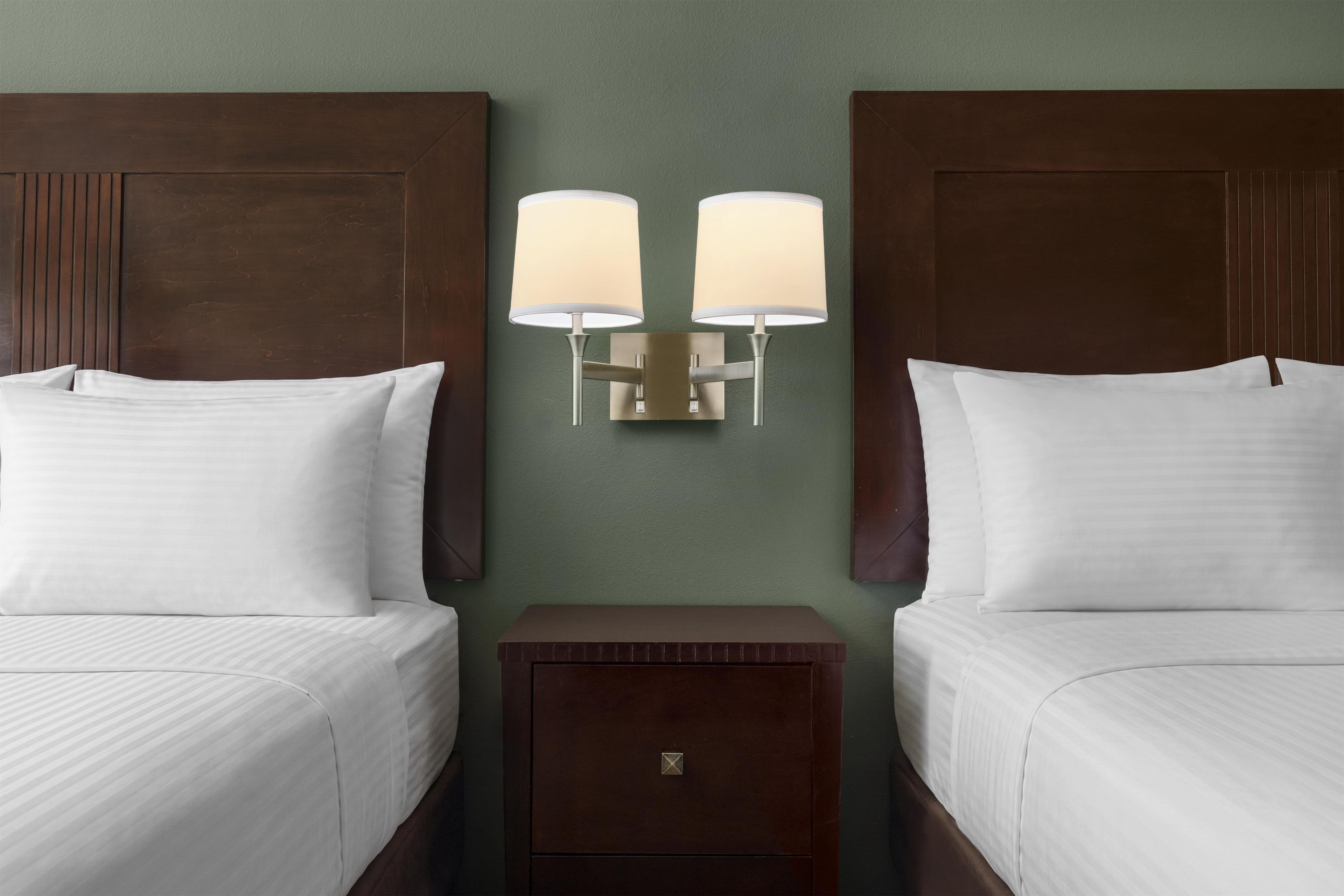 A hotel room with two beds, a nightstand, and a wall-mounted lamp between them.