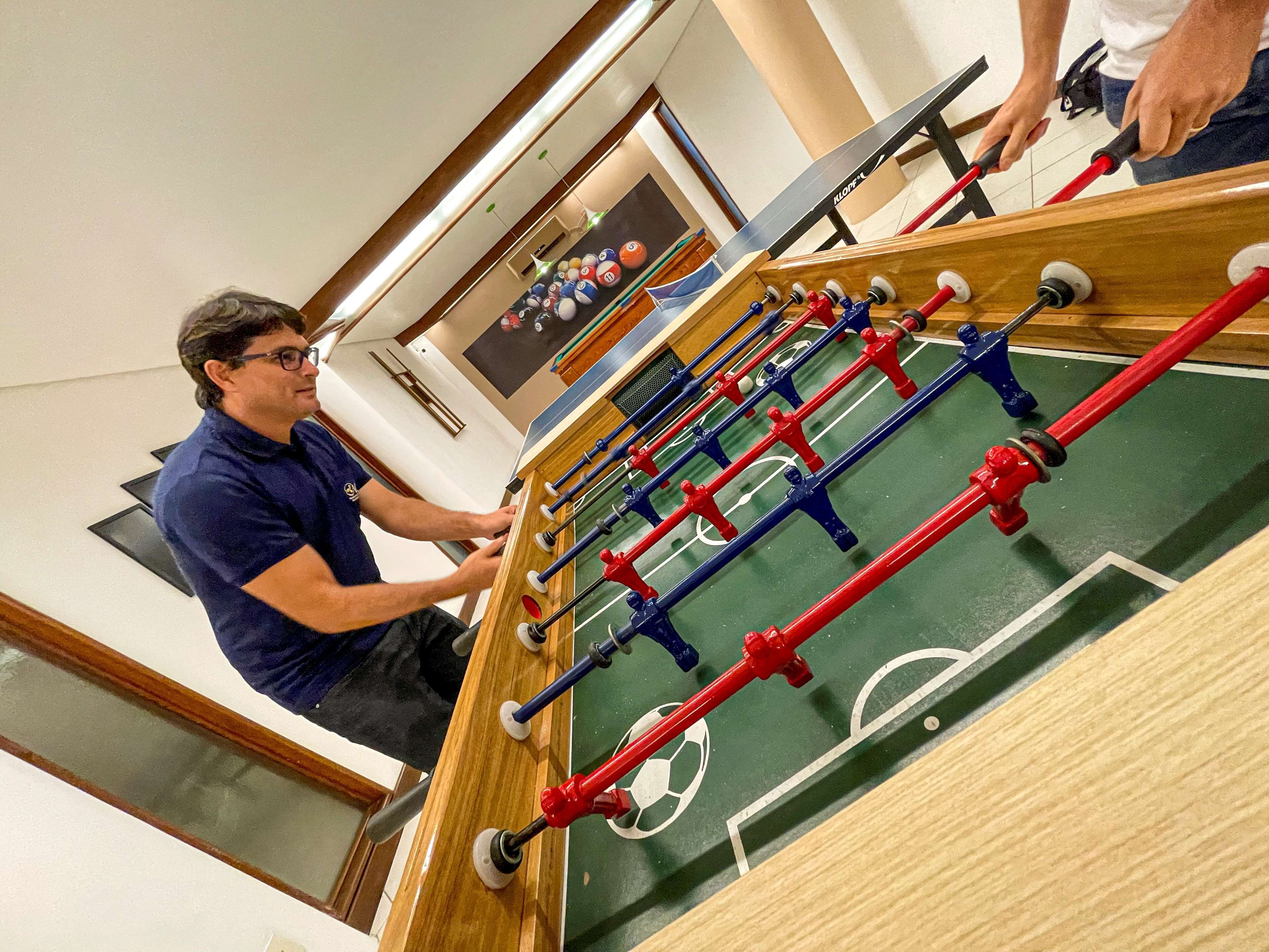 Uma mesa de futebol de mesa com design de campo de futebol, uma pessoa jogando e uma mesa de bilhar ao fundo.