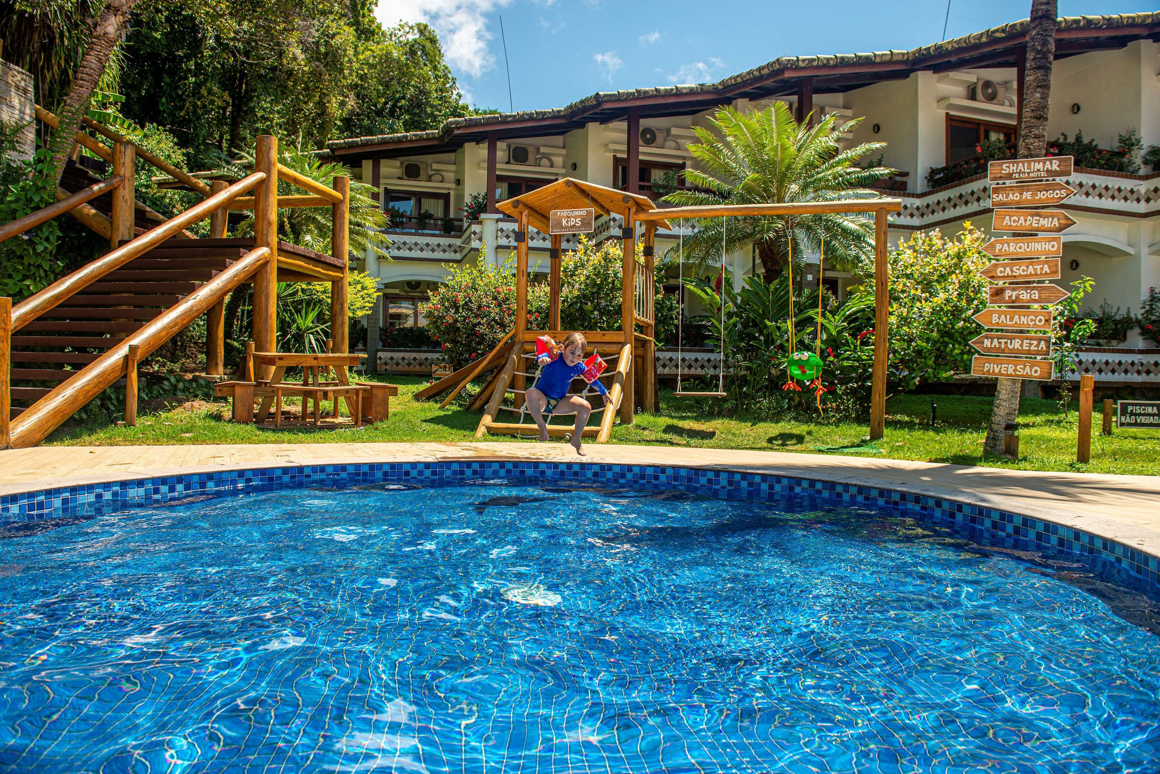 Ein Poolbereich mit einer Holzrutsche und einem Hinweisschild, das Annehmlichkeiten wie 'Shalimar', 'Salão de Jogos' und 'Piscina' kennzeichnet.