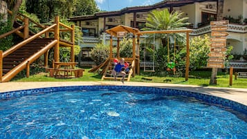 Ein Poolbereich mit einer Holzrutsche und einem Hinweisschild, das Annehmlichkeiten wie 'Shalimar', 'Salão de Jogos' und 'Piscina' kennzeichnet.