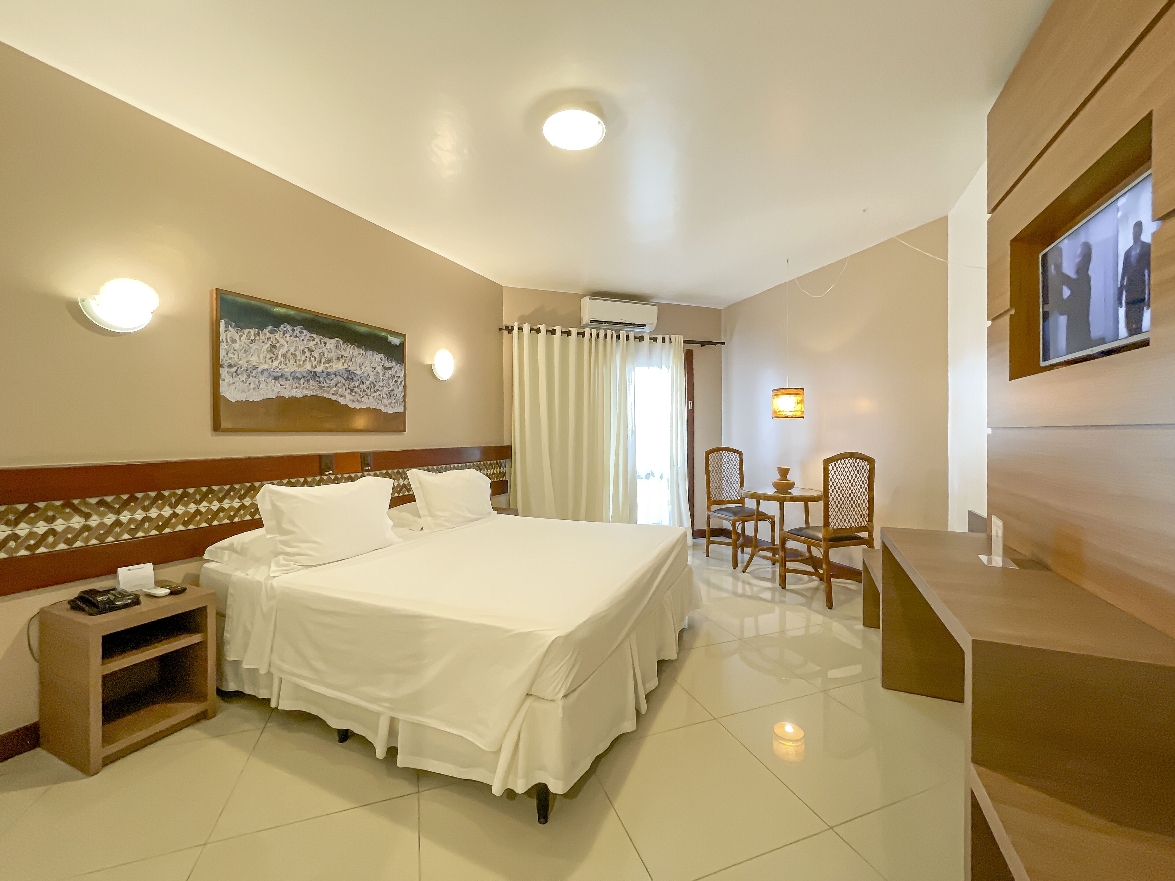 A hotel room with a large bed, a desk with chairs, a painting on the wall, and a television mounted on the wall.