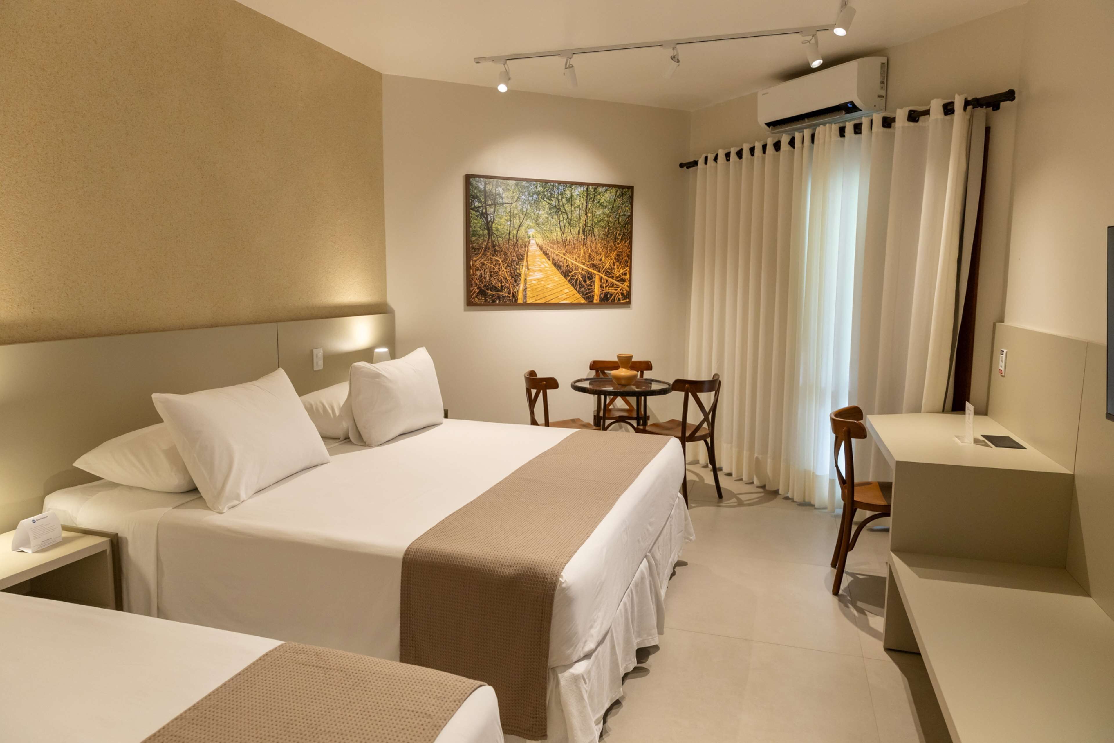 Une chambre d’hôtel avec un grand lit, une table de chevet avec une lampe, une petite table ronde avec des chaises, un bureau avec une chaise et un tableau accroché au mur.
