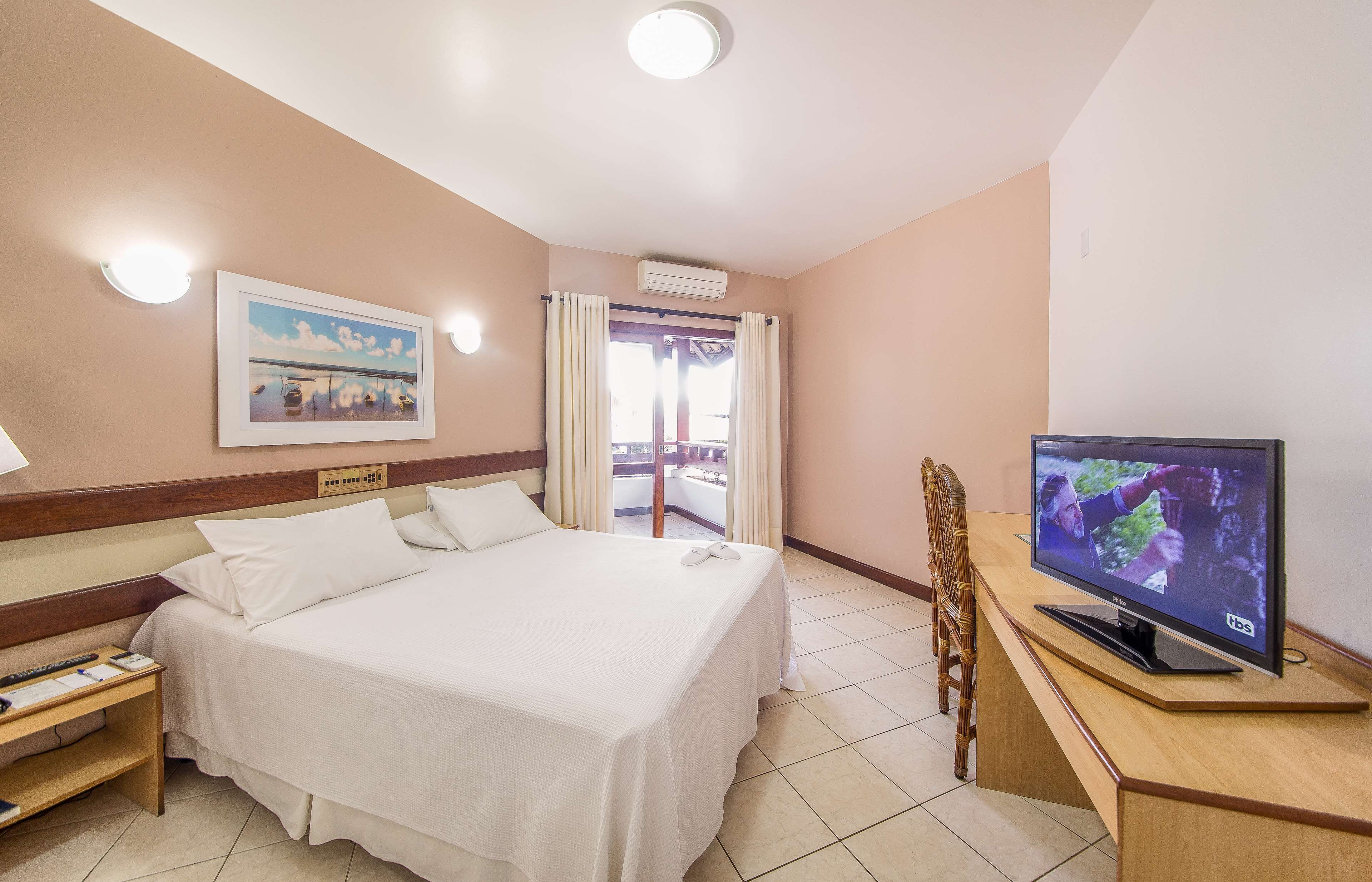 A hotel room with a bed, a desk with a TV, a chair, a nightstand, and a window with curtains.