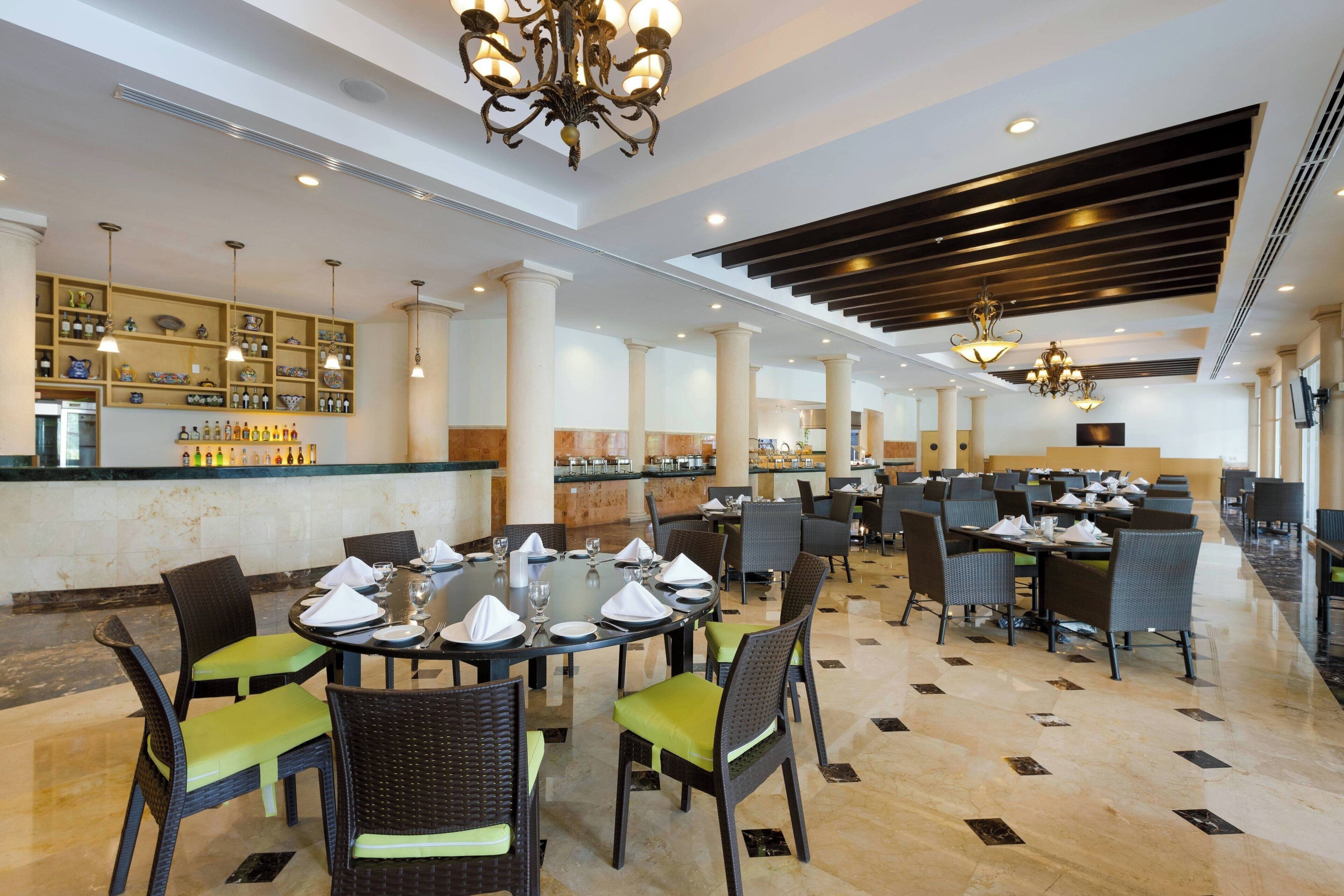 A spacious dining area with round tables, green cushioned chairs, and a bar area with shelves of bottles.