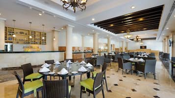 A spacious dining area with round tables, green cushioned chairs, and a bar area with shelves of bottles.