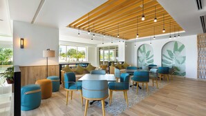 Un intérieur moderne avec des poutres en bois au plafond, des sièges bleus et orange, et de grandes fenêtres offrant une vue sur la verdure.