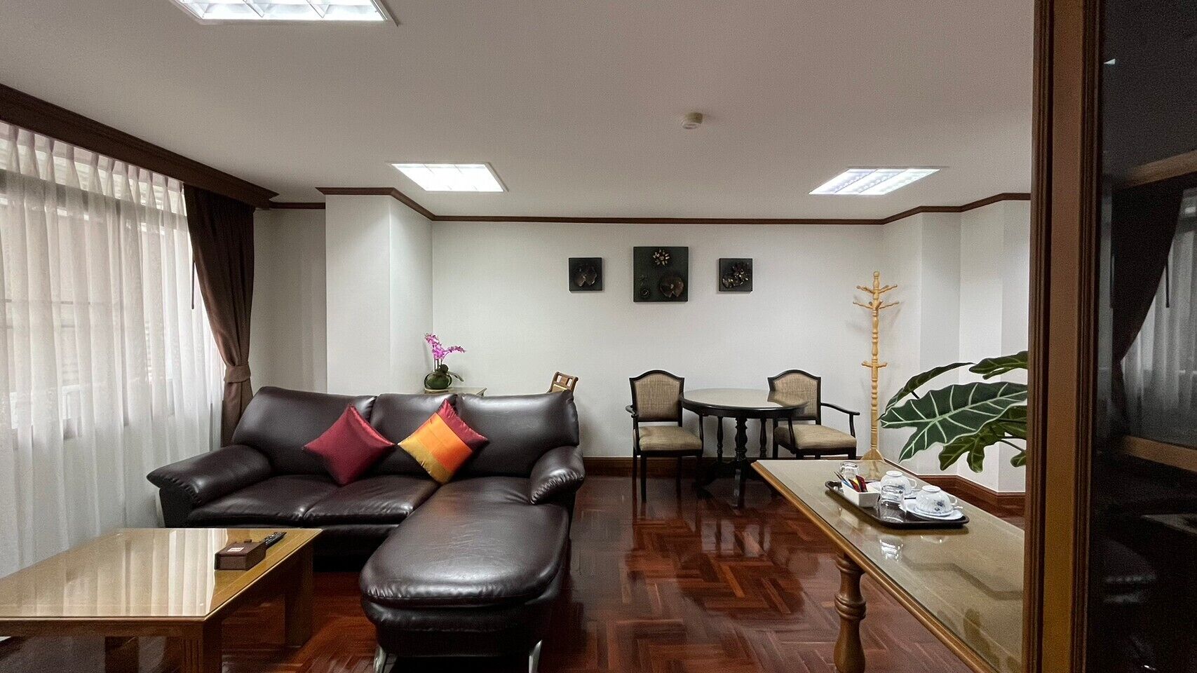 Ein modernes Wohnzimmer mit einem Ledersofa, einem kleinen Esstisch und einem Couchtisch. Eine große Pflanze und eine Uhr sind an der Wand zu sehen.