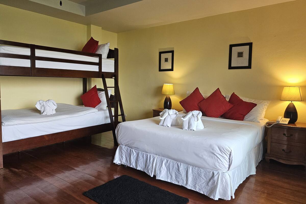 A hotel room with two bunk beds, a large bed, wooden flooring, two bedside tables with lamps, and a black rug.