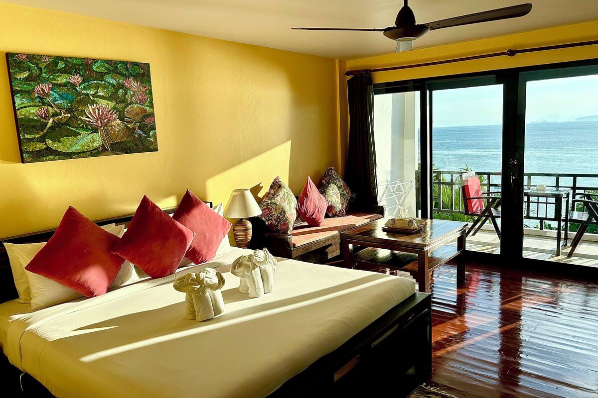 A bedroom with a large bed, a ceiling fan, a painting of water lilies, and a balcony with a view of the sea.